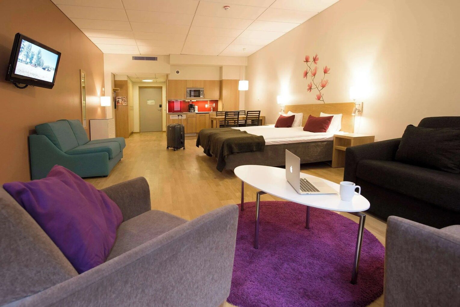 A hotel room with a bed, a desk with a laptop, a TV, a sofa, and a small kitchenette.