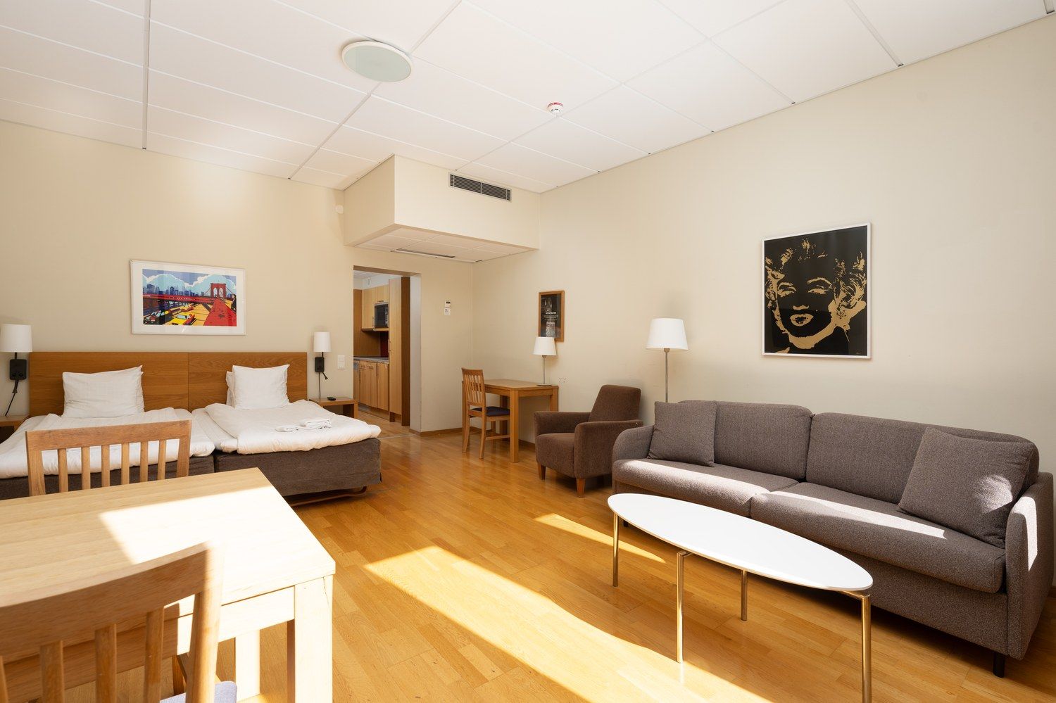 Ett hotellrum med en säng, en soffa, ett matbord och en stol. Man kan se ett köksutrymme genom en öppen dörr.