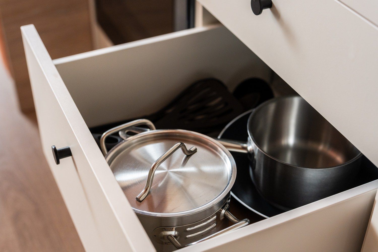 Un tiroir contenant une casserole en acier inoxydable avec un couvercle, une petite casserole noire et une paire de cuillères en bois.