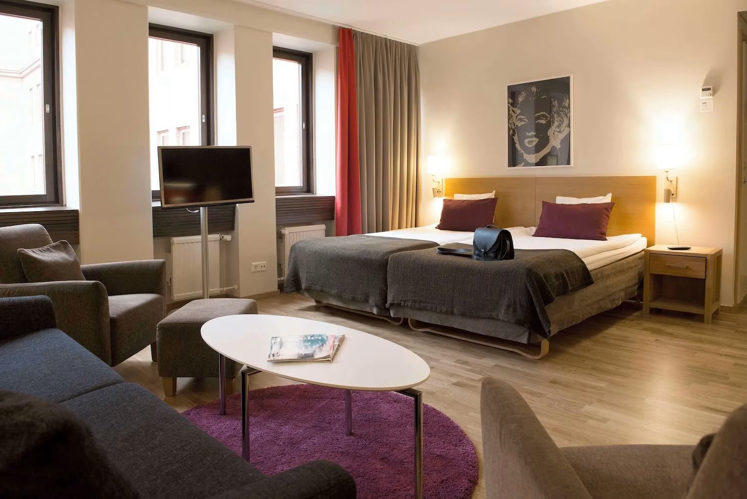 Habitación de hotel con cama, dos mesitas de noche, televisor, sofá, una mesita pequeña y ventana con cortinas.