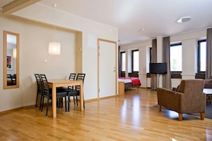 Una habitación de hotel moderna con una mesa de comedor de madera, sillas, una cama con una colcha roja, un televisor y ventanas grandes.