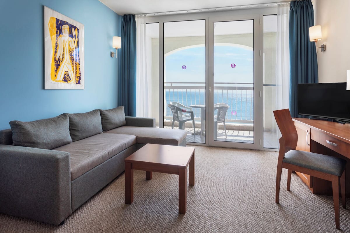 A hotel room with a grey sofa, a wooden coffee table, a TV stand with a TV, and a balcony with a view of the sea.