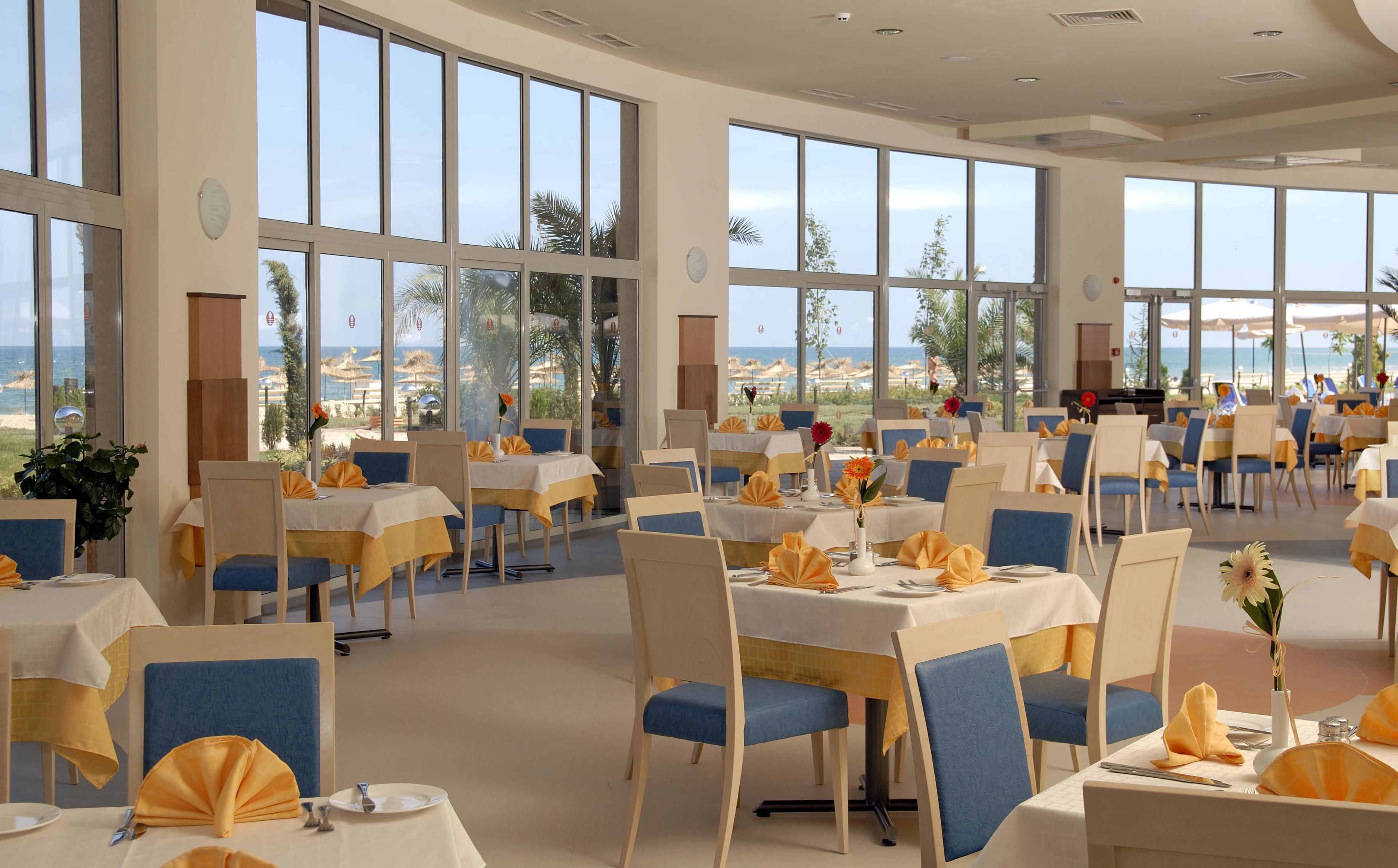 Un comedor amplio con ventanas grandes que ofrecen vistas a la playa y al océano. Las mesas están ordenadas con manteles amarillos y azules, y fundas de silla a juego.