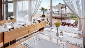 A restaurant with wooden benches and tables, white tablecloths, and a view of the beach and boats. - Sol Luna Bay Resort (Byala)