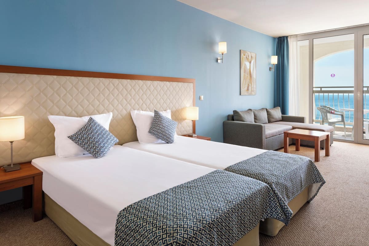 A hotel room with two beds, a sofa, a balcony with a view of the sea, and a large window.