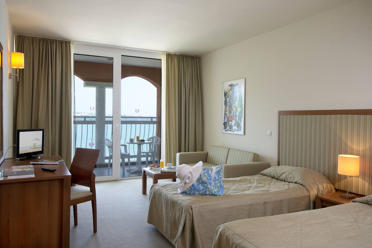 A hotel room with a desk, two beds, a chair, a table, and a balcony with a view.