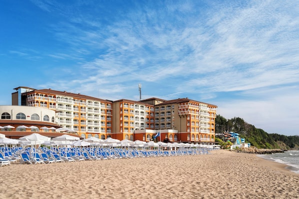 A beachfront hotel with multiple floors, a sandy beach with blue lounge chairs, and umbrellas. - Sol Luna Bay Resort (Byala)
