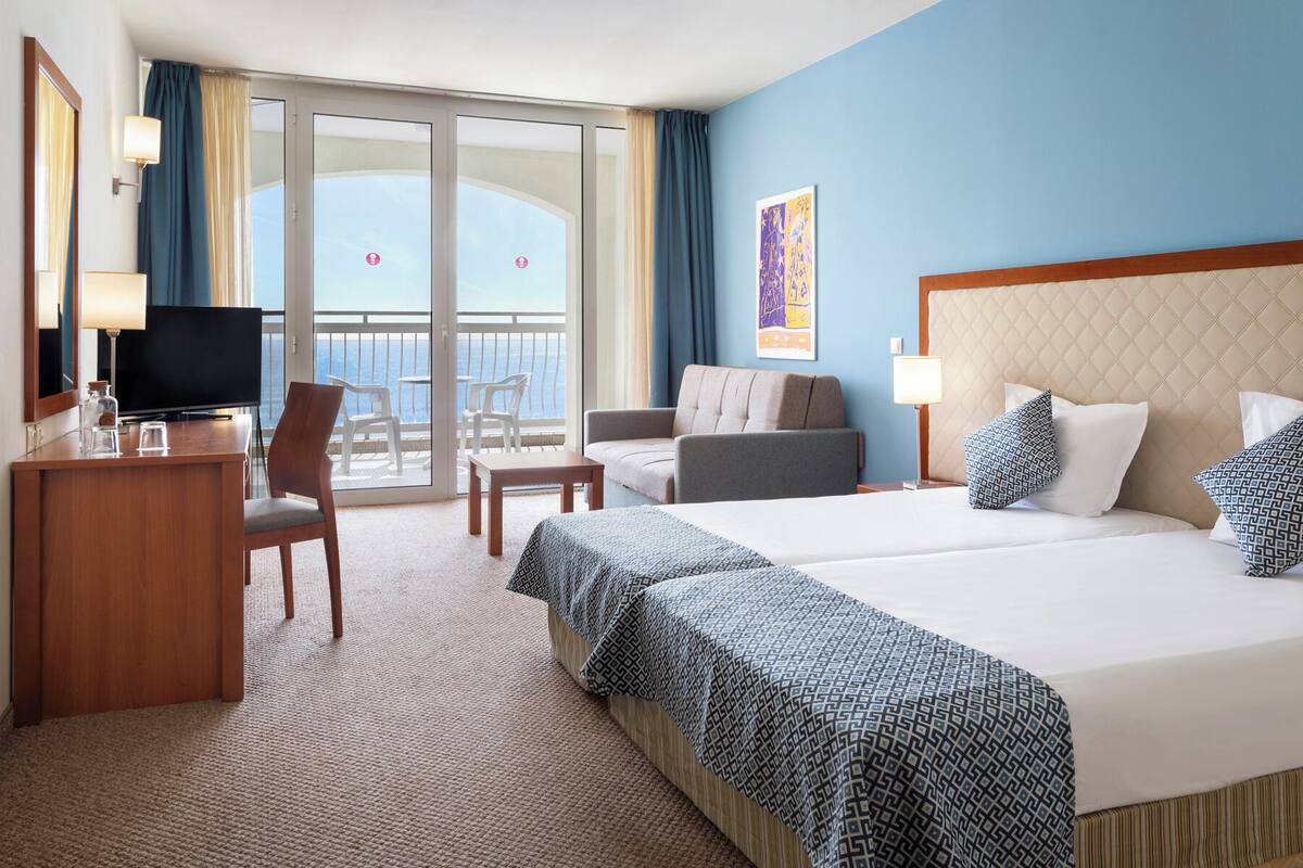 A hotel room with a bed, a desk, a TV, and a balcony with a view of the sea.