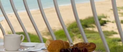 Een tafel met een boek, een kopje, een bord met croissantjes en kersen, en een vork, met uitzicht op een strand.