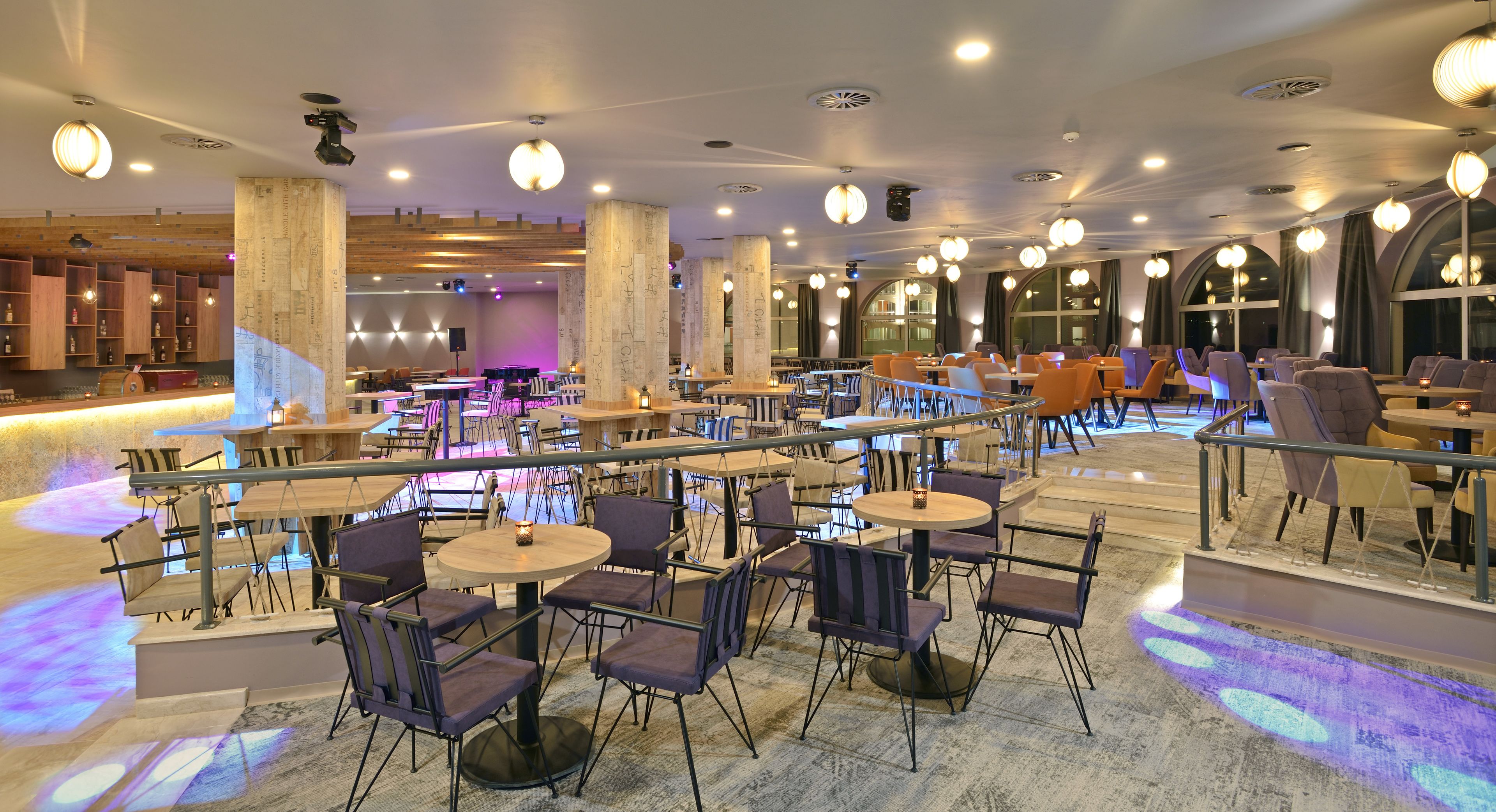 Un'ampia sala da pranzo interna con diversi tavoli e sedie, ampie finestre e moderni corpi illuminanti.