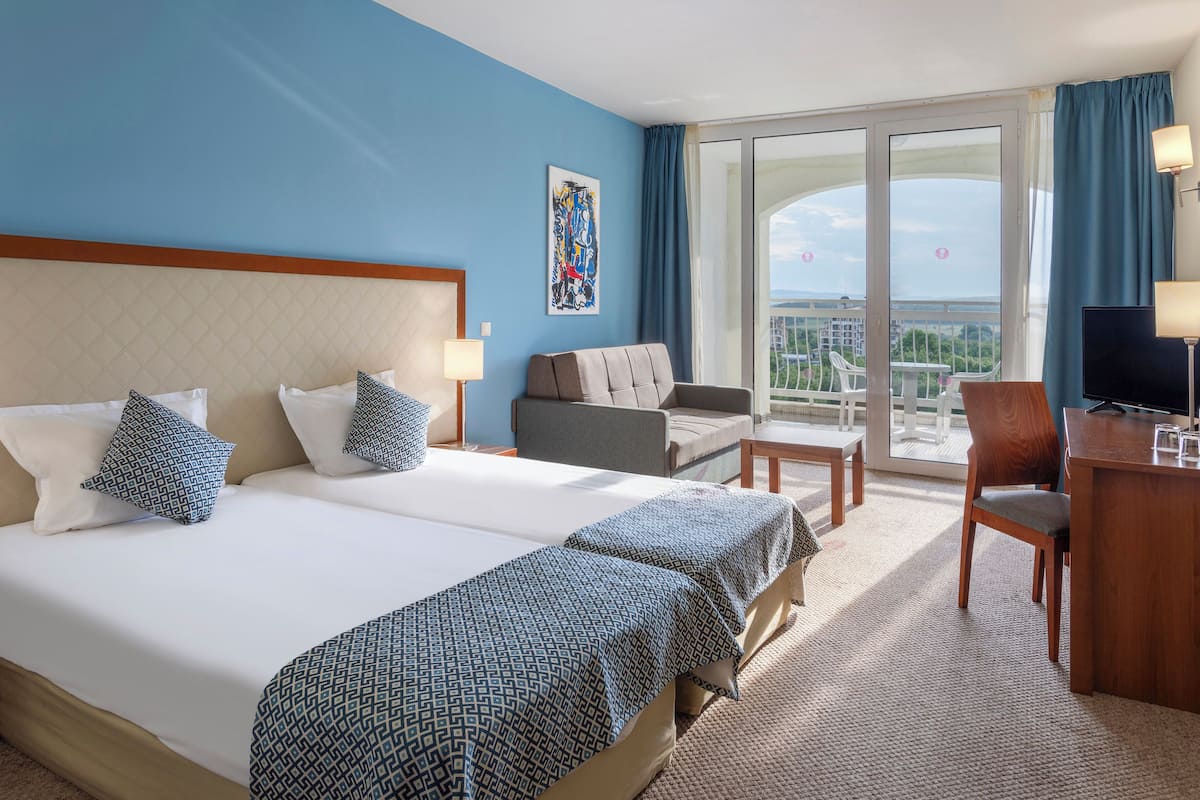 A hotel room with two beds, a desk, a chair, and a balcony with a view.
