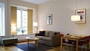 Ein Wohnzimmer mit Sofa, einem kleinen runden Tisch, einem Esstisch mit Stühlen und einem Fenster mit Vorhängen.