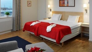 Ein Hotelzimmer mit einem Bett, zwei Nachttischen, einem Fenster mit Vorhängen, einer Topfpflanze und Blick auf Gebäude.