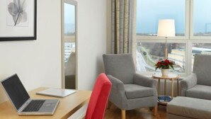 Ein Hotelzimmer mit einem Holztisch, einem Laptop, einem roten Sessel, zwei grauen Sesseln, einem kleinen Tisch mit einer Lampe und einem Fenster mit Stadtblick.