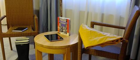 Una silla de mimbre con un cojín amarillo, una mesa redonda de madera con un libro y una tableta, y un cubo de basura con la etiqueta 'AIGLE' en una habitación con cortinas.