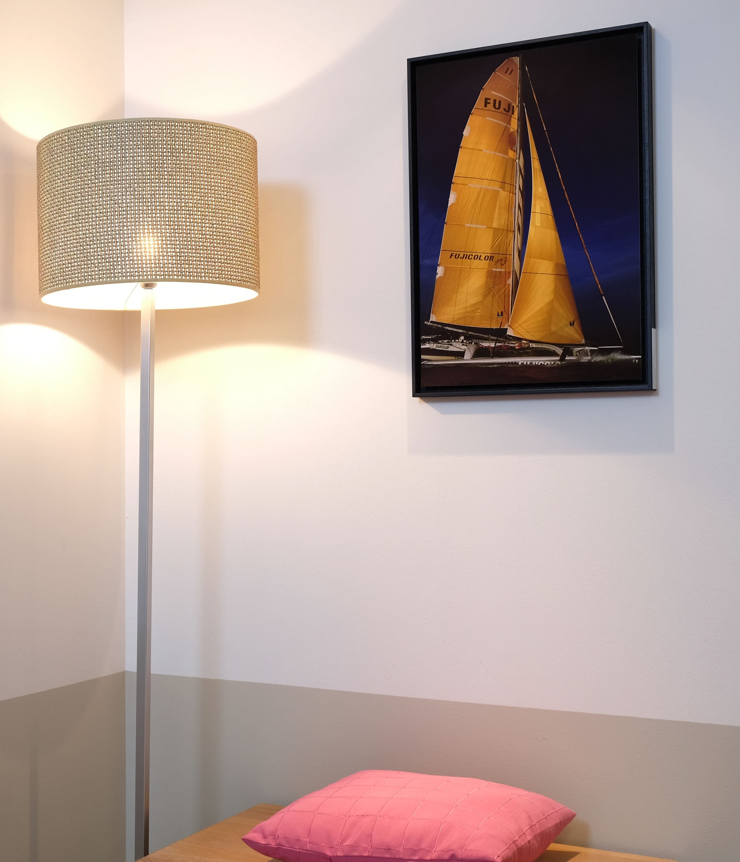 Una habitación con una lámpara de pie, un cuadro con una imagen de un velero y un cojín rosa sobre una superficie de madera.