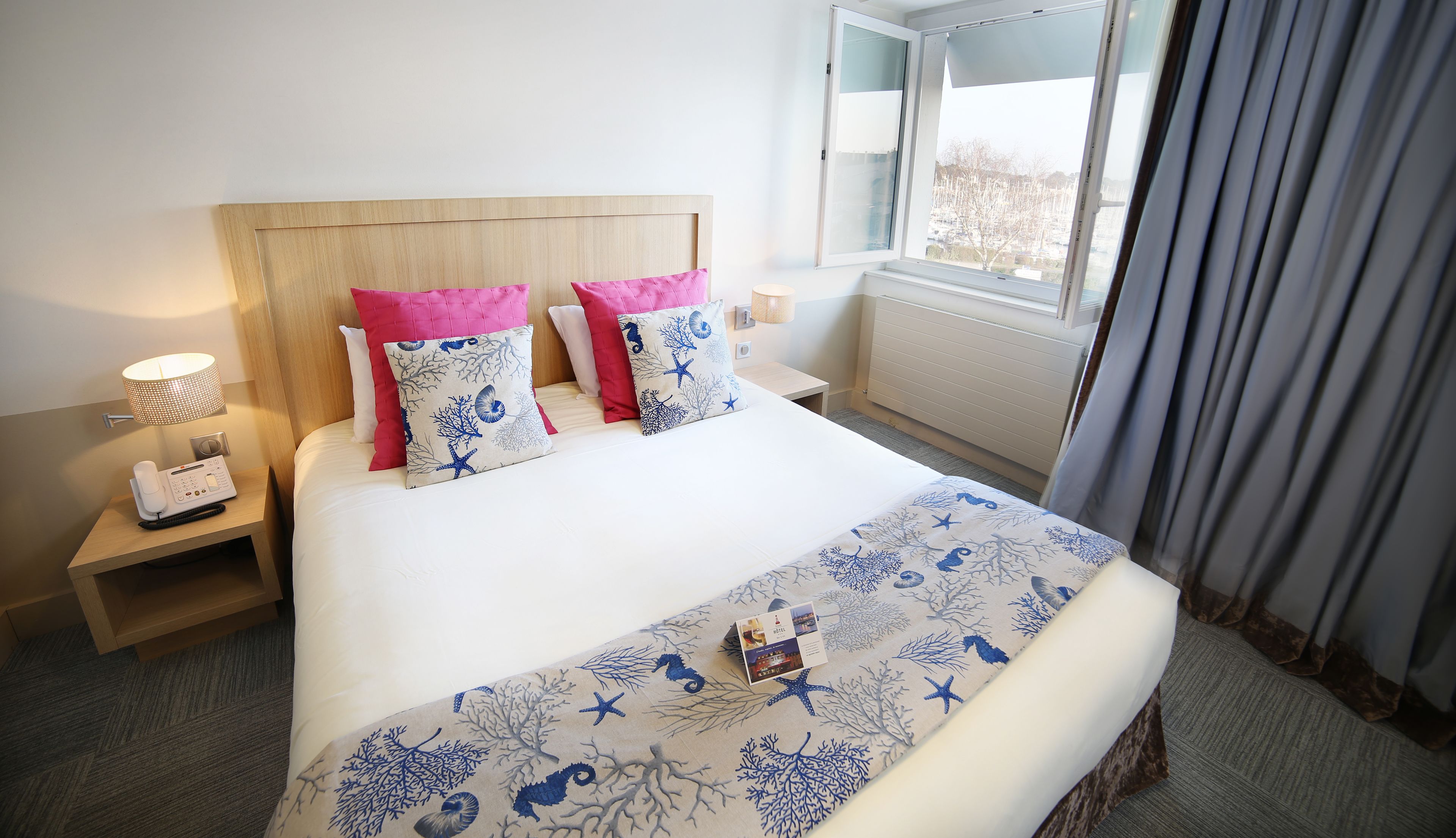 Een hotelkamer met een bed, nachtkastje met telefoon, lamp en raam met gordijnen.