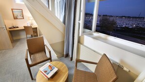 Habitación de hotel con balcón, un escritorio, dos sillas de mimbre y vistas a un puerto deportivo al atardecer.