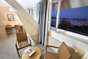 A hotel room with a balcony, a desk, two wicker chairs, and a view of a marina at dusk.