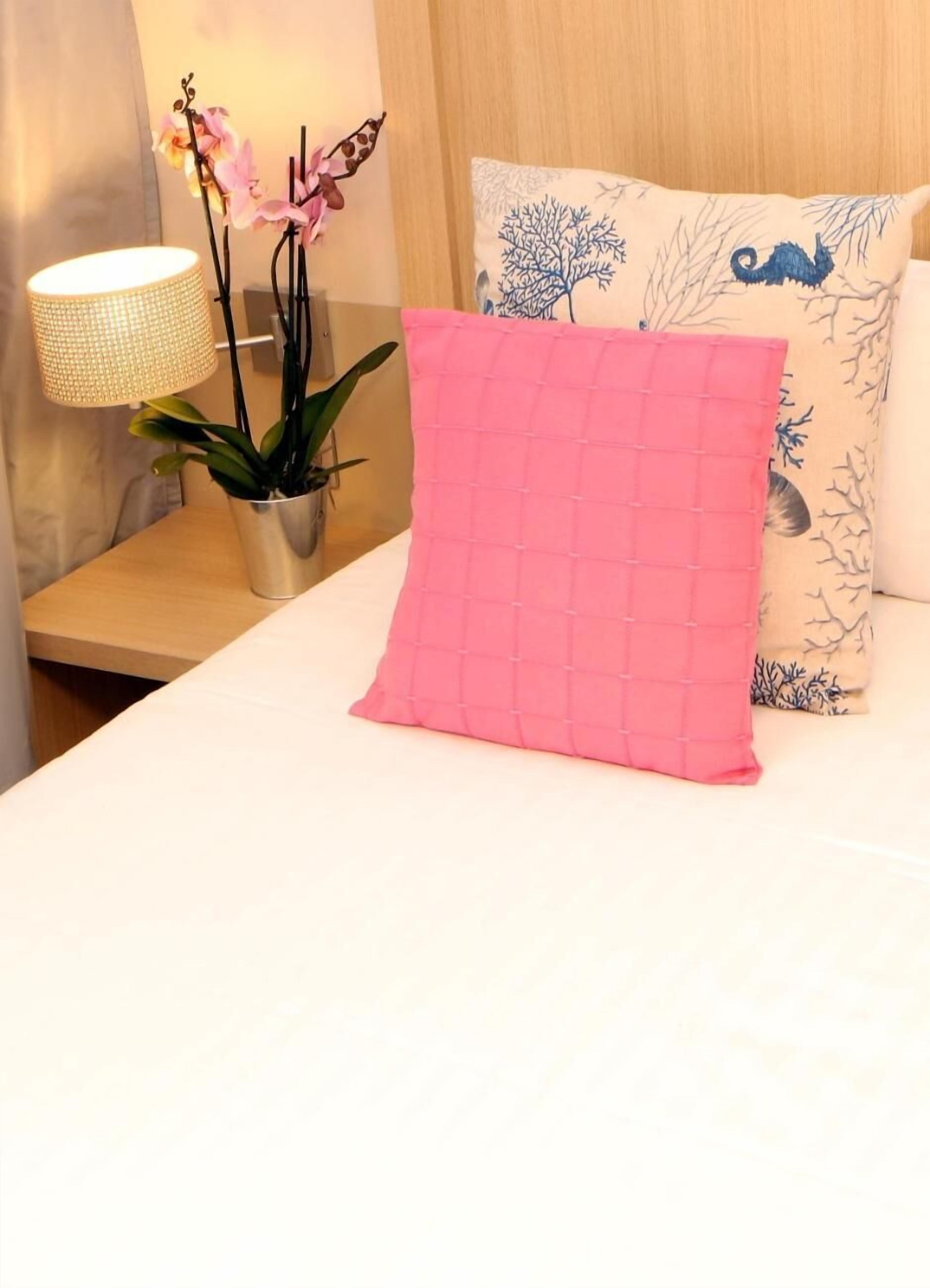 A neatly made bed with a pink quilted pillow, a bedside lamp, and a potted plant with pink flowers.