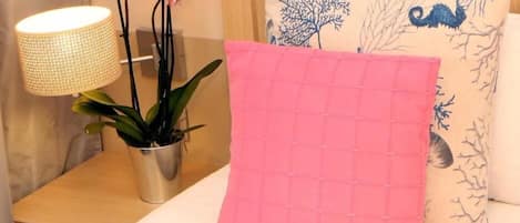A neatly made bed with a pink quilted pillow, a bedside lamp, and a potted plant with pink flowers.