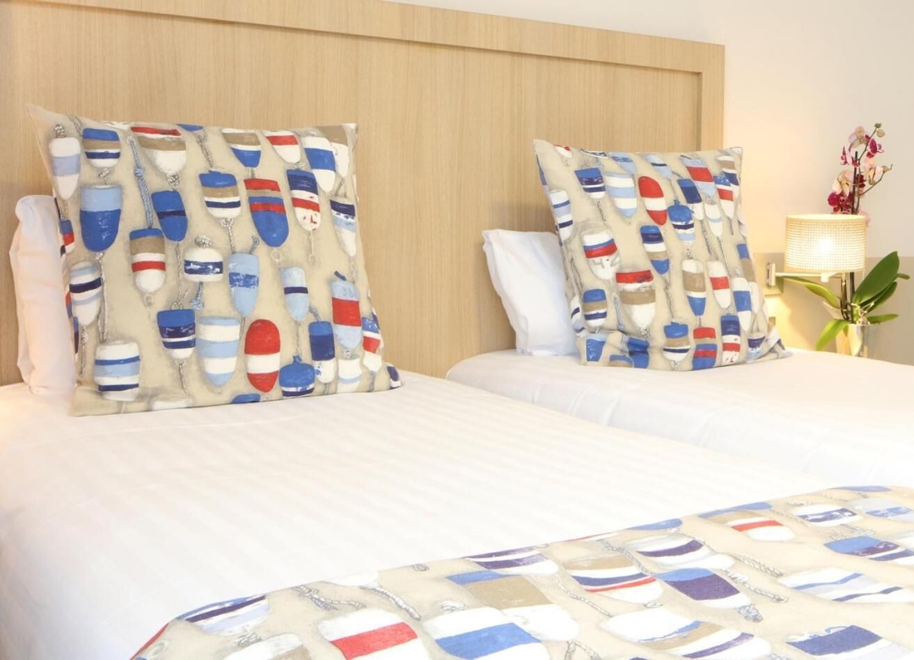 Une chambre d’hôtel avec un grand lit, deux oreillers à motifs nautiques, une table de chevet avec une lampe et une plante en pot.