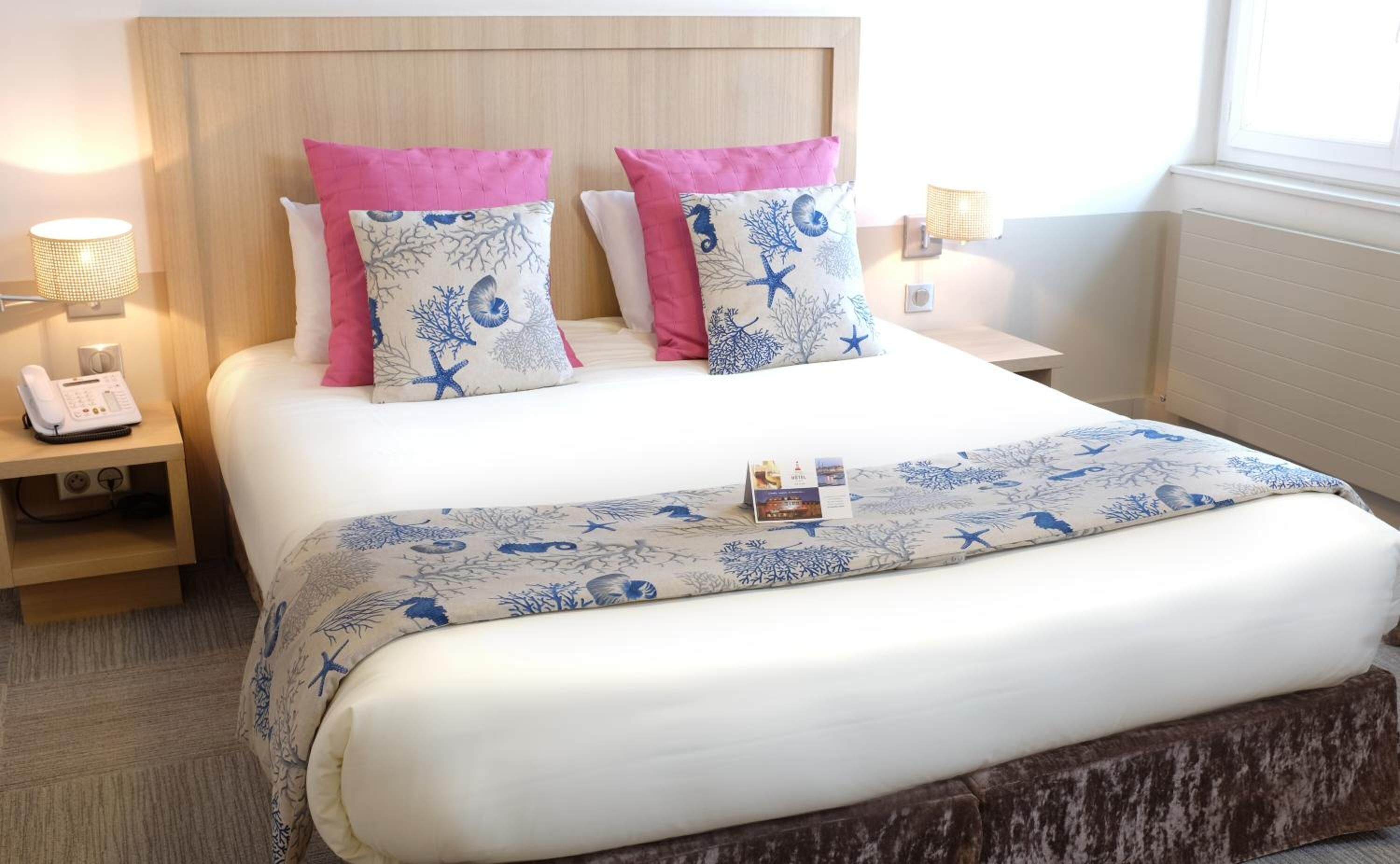 Un lit bien fait, recouvert d’une couvre-lit à motifs floraux blancs et bleus, agrémenté de deux coussins roses et doté d’une tête de lit en bois.