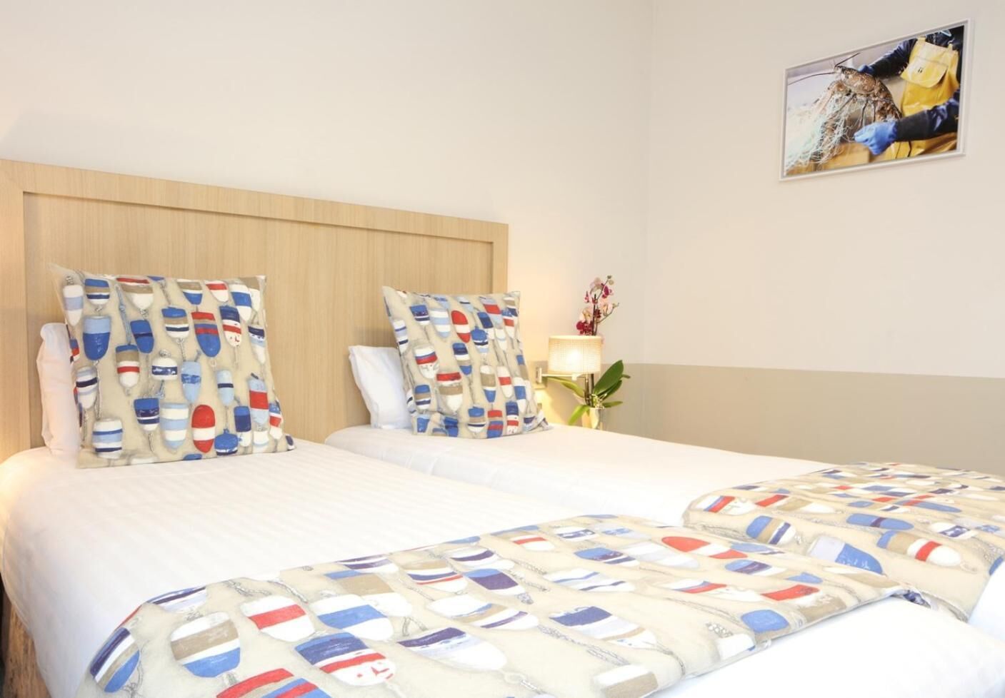A double bed with a wooden headboard, two pillows with nautical patterns, and a bedside table with a lamp and a plant.