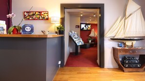 Un área de recepción con un escritorio de madera, una pequeña decoración de velero, un panel de comentarios de huéspedes y un pasillo con alfombra roja.