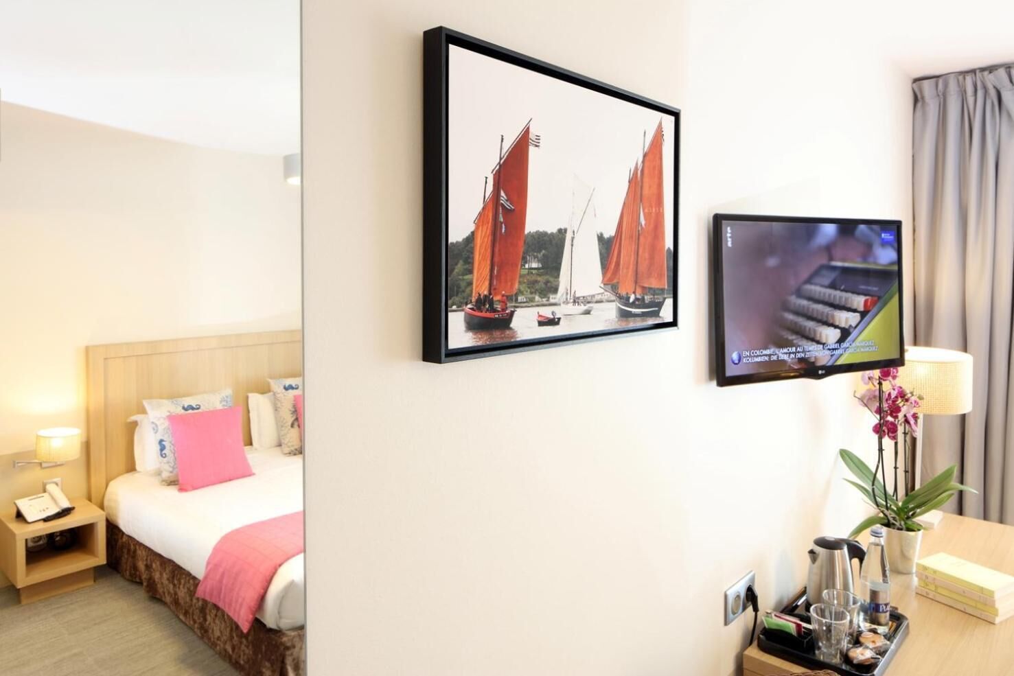 Une chambre d’hôtel avec un lit, une télévision, un tableau encadré et un bureau avec une lampe et une plante.