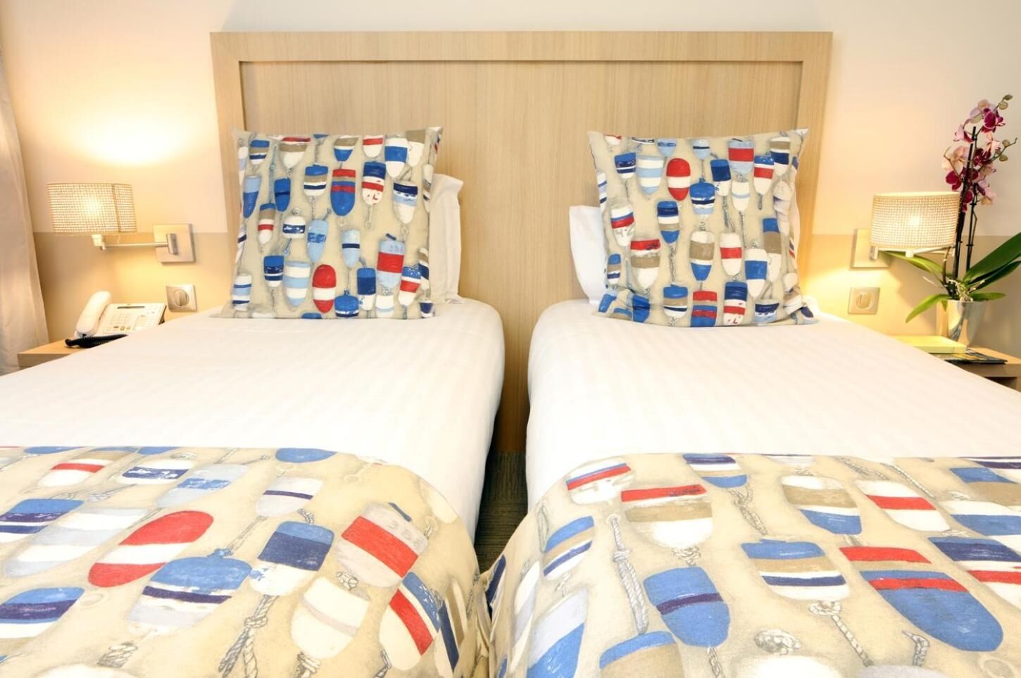 Ein Hotelzimmer mit zwei Betten, jeweils mit einem gemusterten Bettbezug und passenden Kissen. Ein Nachttisch beherbergt eine Lampe, ein Telefon und eine kleine Pflanze. Das Zimmer verfügt über einen Holz-Kopfteil und eine Wandlampe.