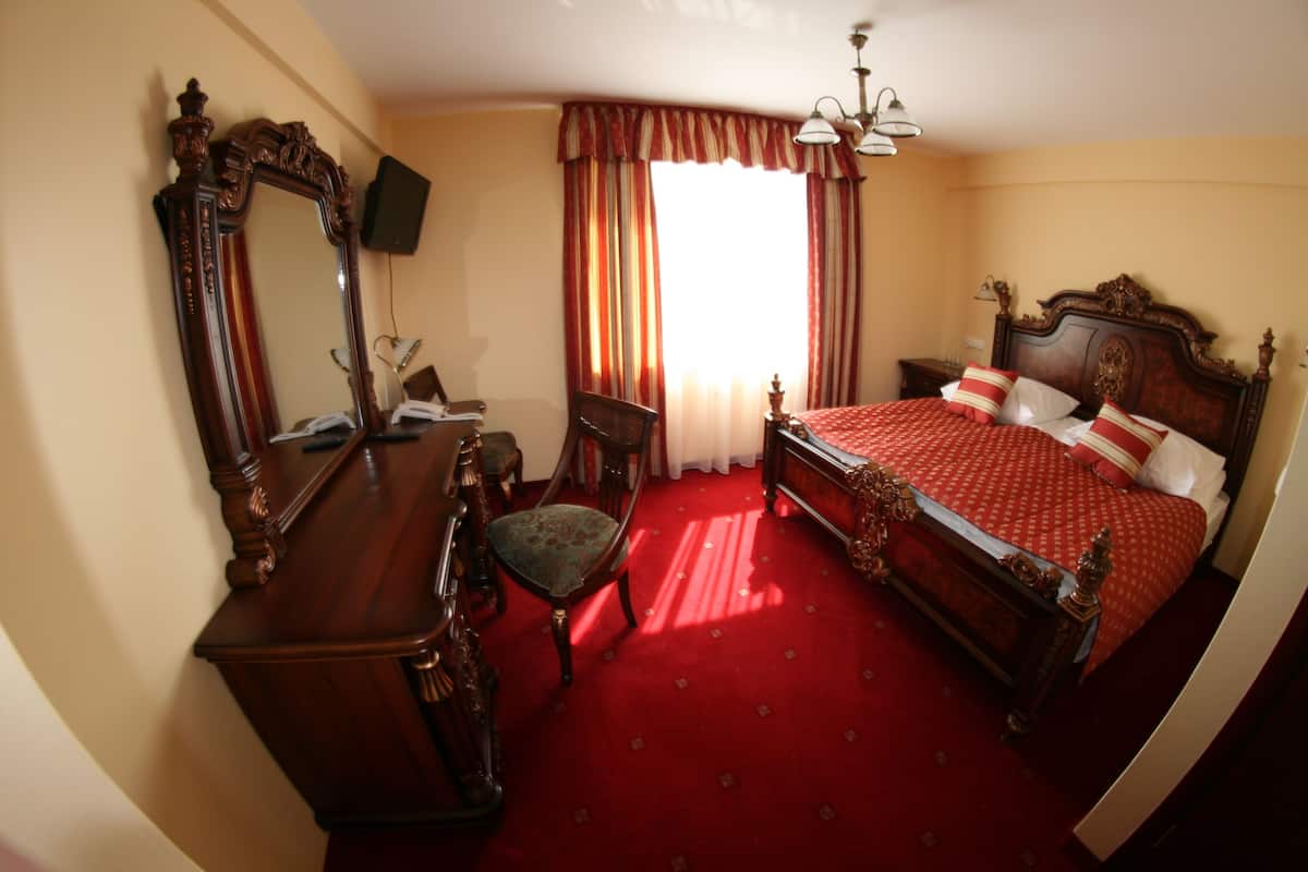 A hotel room with a large ornate bed, a wooden dresser, a chair, and a television on the wall.