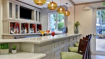 Zona de bar con barra, varias botellas de licor y una pantalla de televisión. Hay sillas con cojines y una ventana con vistas al exterior.