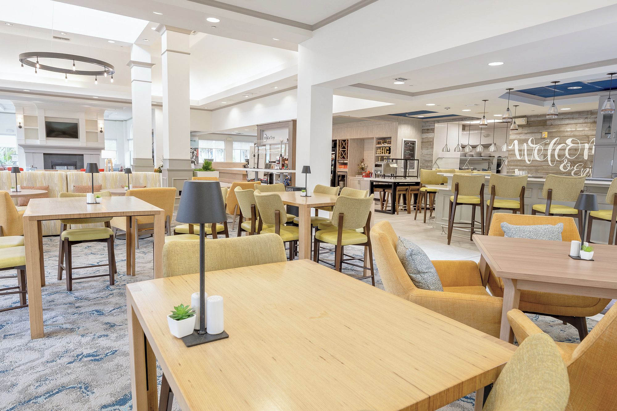 A spacious hotel lobby with multiple seating areas, a fireplace, and a modern kitchen visible in the background.