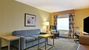Habitación de hotel con sofá, una mesita, una silla, escritorio, televisor y ventana con cortinas.
