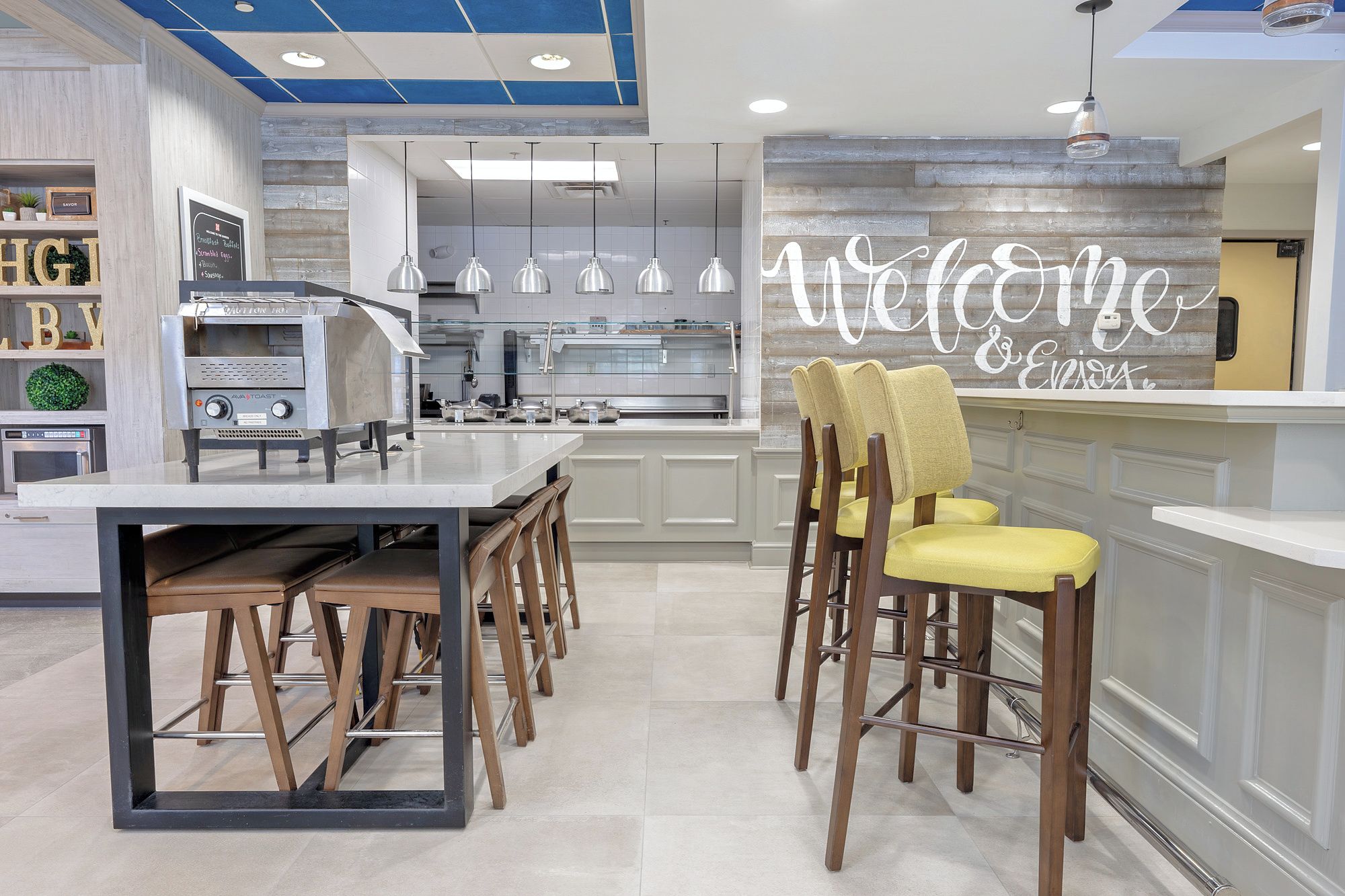 A modern kitchen with a built-in oven, a coffee machine, and a bar area with a 'Welcome & Enjoy' sign.