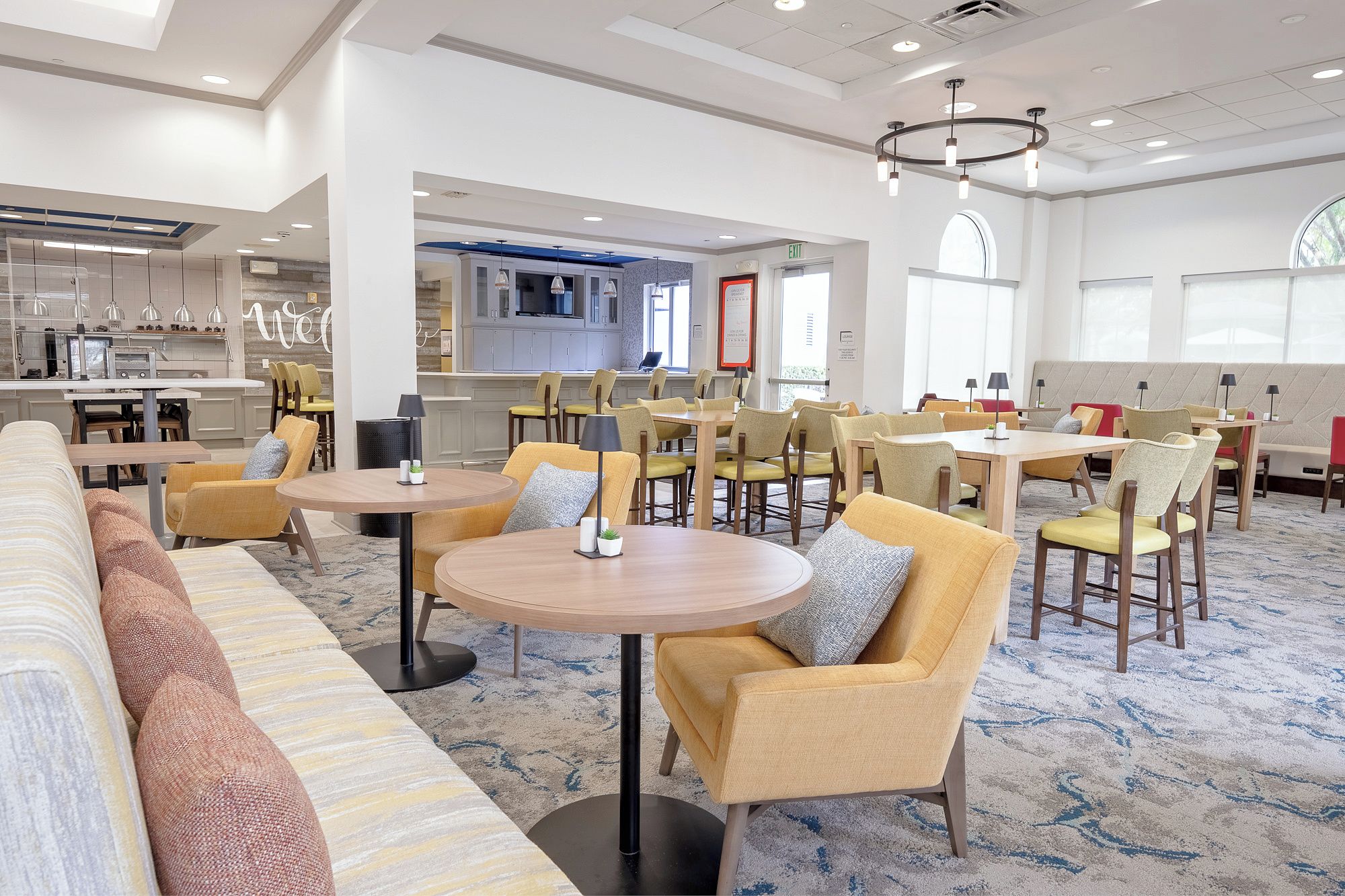 A hotel lobby with a dining area, sofas, and a kitchen in the background.