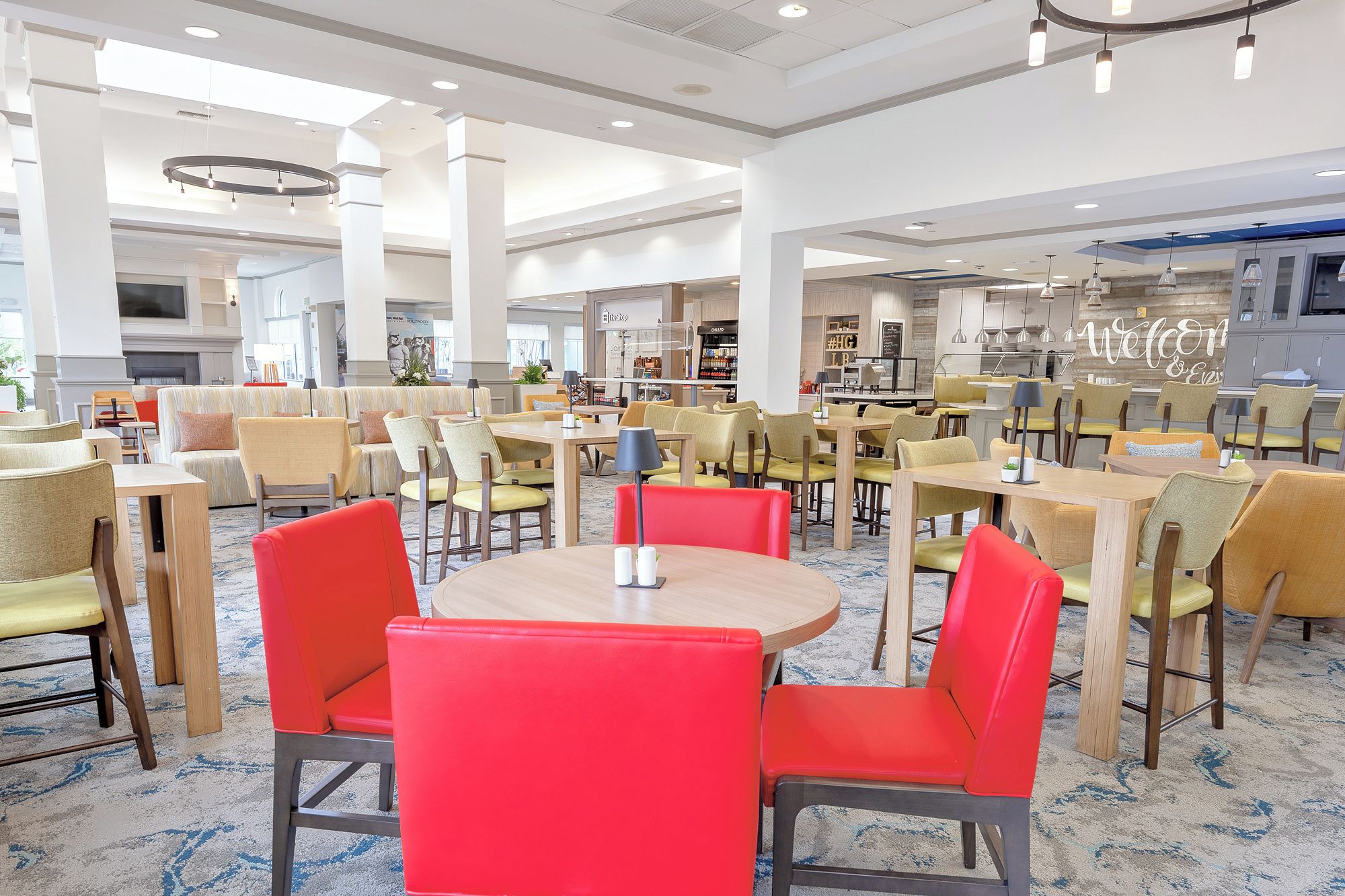 Un hall de hotel con varias zonas de estar, que incluyen mesas y sillas redondas, y un área de restaurante visible al fondo.