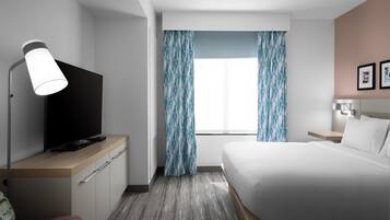 A hotel room with a bed, a TV on a wooden dresser, a desk with a lamp, a window with curtains, and a bedside table with a lamp.