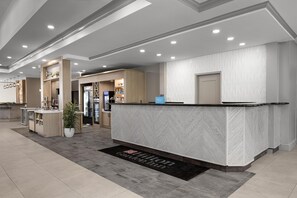 Zona de recepción de hotel con mostrador de mármol, una mesa de recepción, una planta e indicación del nombre del hotel.