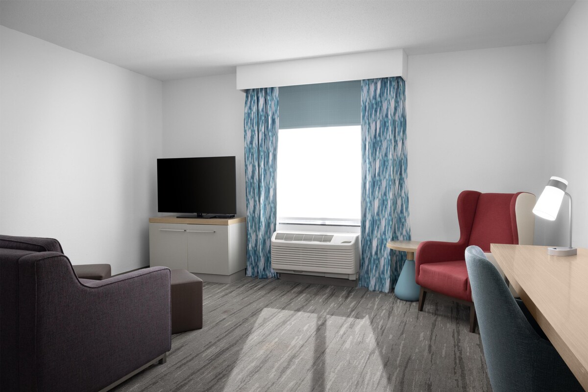 Une chambre d’hôtel équipée d’un téléviseur à écran plat, d’un bureau avec une lampe, d’un fauteuil rouge et d’un canapé gris.