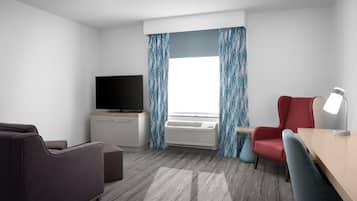 Habitación de hotel con televisión de pantalla plana, escritorio con lámpara, una silla roja y un sofá gris.