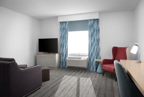 A hotel room with a flat-screen TV, a desk with a lamp, a red chair, and a grey sofa.