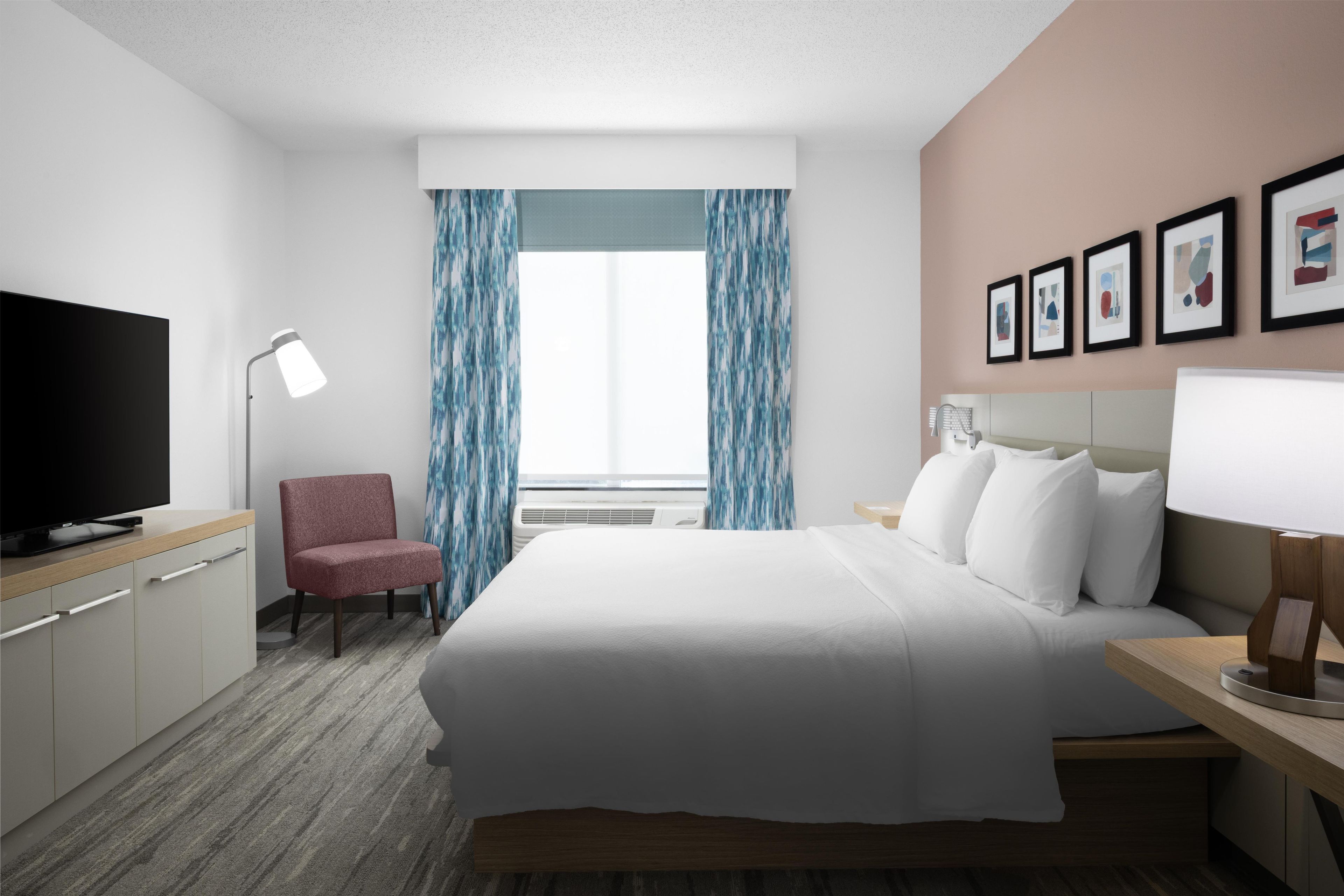 Una habitación de hotel con una cama, una silla, un escritorio con lámpara, televisión y cuadros en la pared.