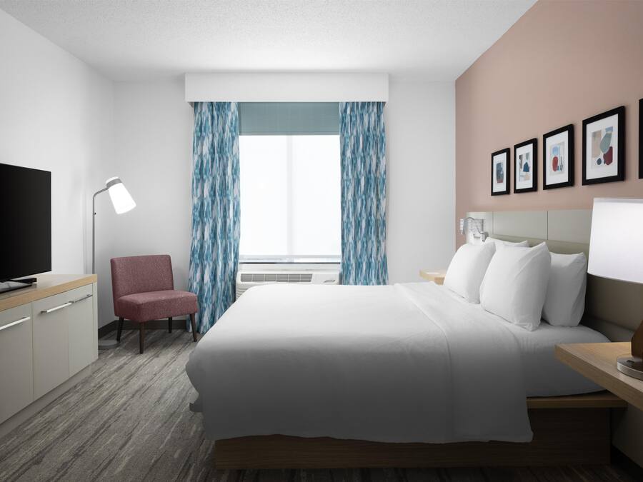 Una habitación de hotel con una cama, una silla, un escritorio con lámpara, televisión y cuadros en la pared.