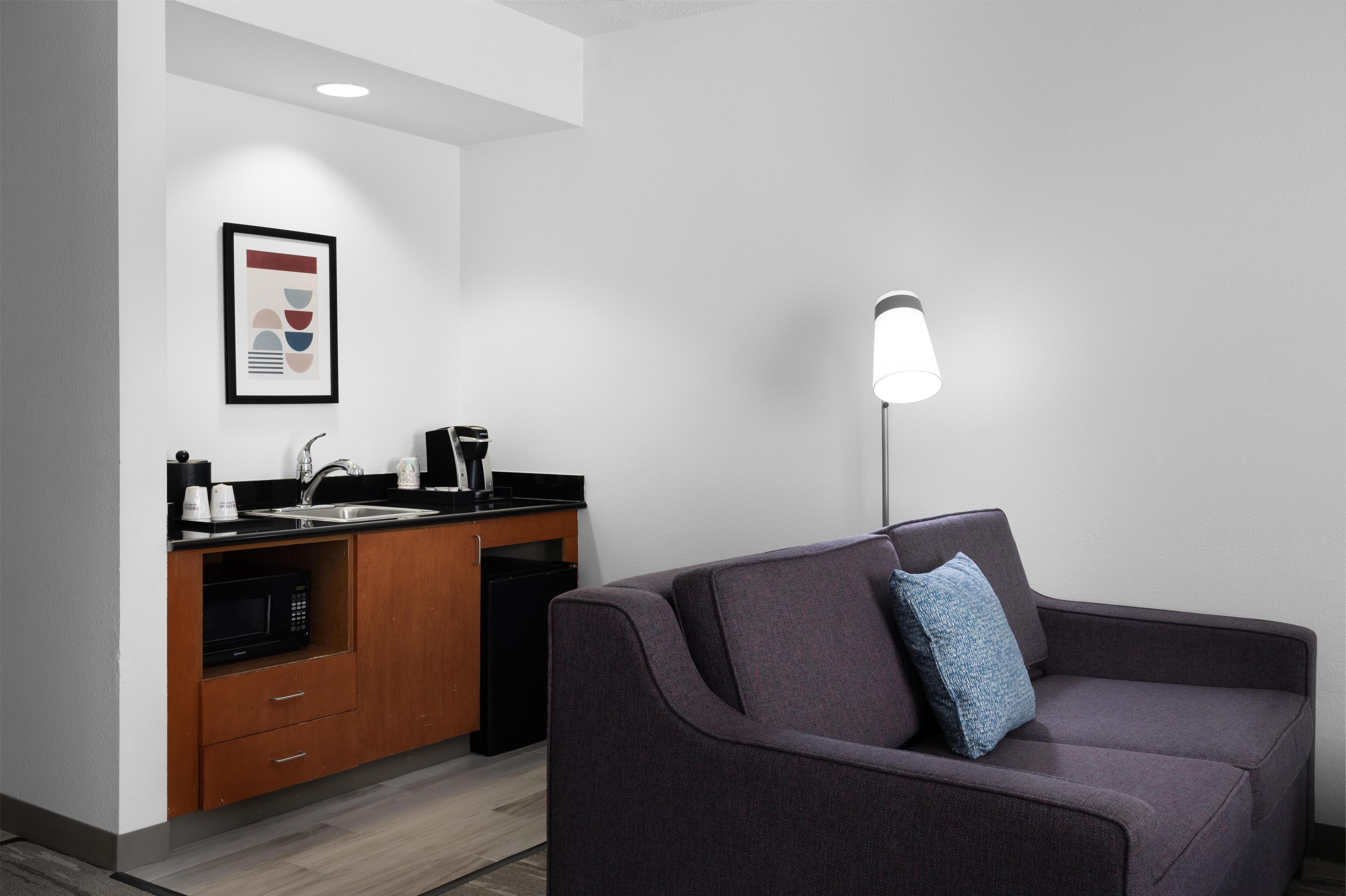 A modern kitchenette with a sink, coffee maker, and microwave, a framed abstract artwork on the wall, and a sofa with a pillow.