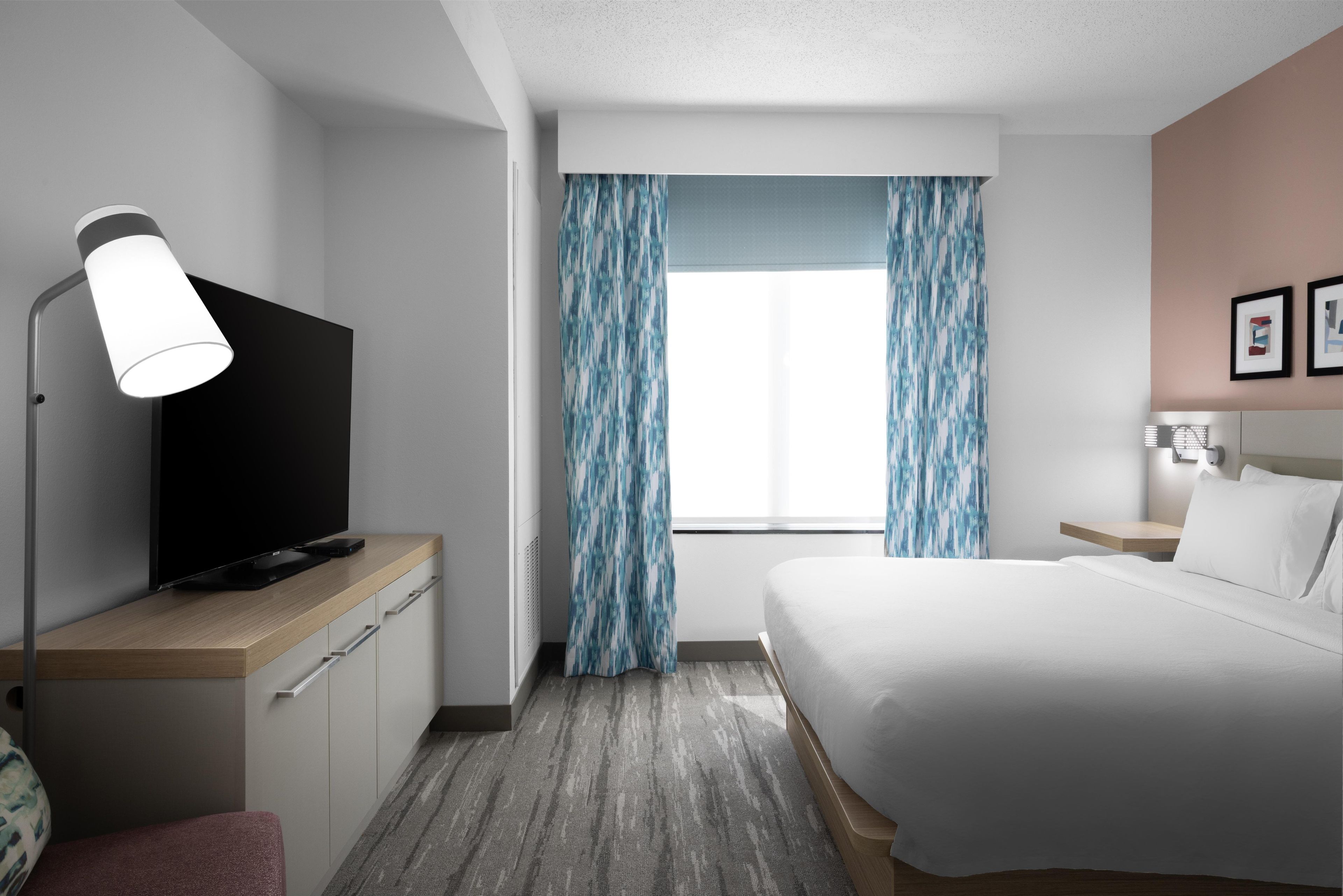 Ein Hotelzimmer mit einem Bett, einem Fernseher auf einer Holzkommode, einem Schreibtisch mit einer Lampe, einem Fenster mit Vorhängen und einem Nachttisch mit einer Lampe.