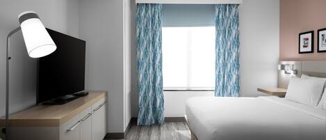 Une chambre d’hôtel avec un lit, un téléviseur posé sur une commode en bois, un bureau avec une lampe, une fenêtre avec des rideaux et une table de chevet avec une lampe.