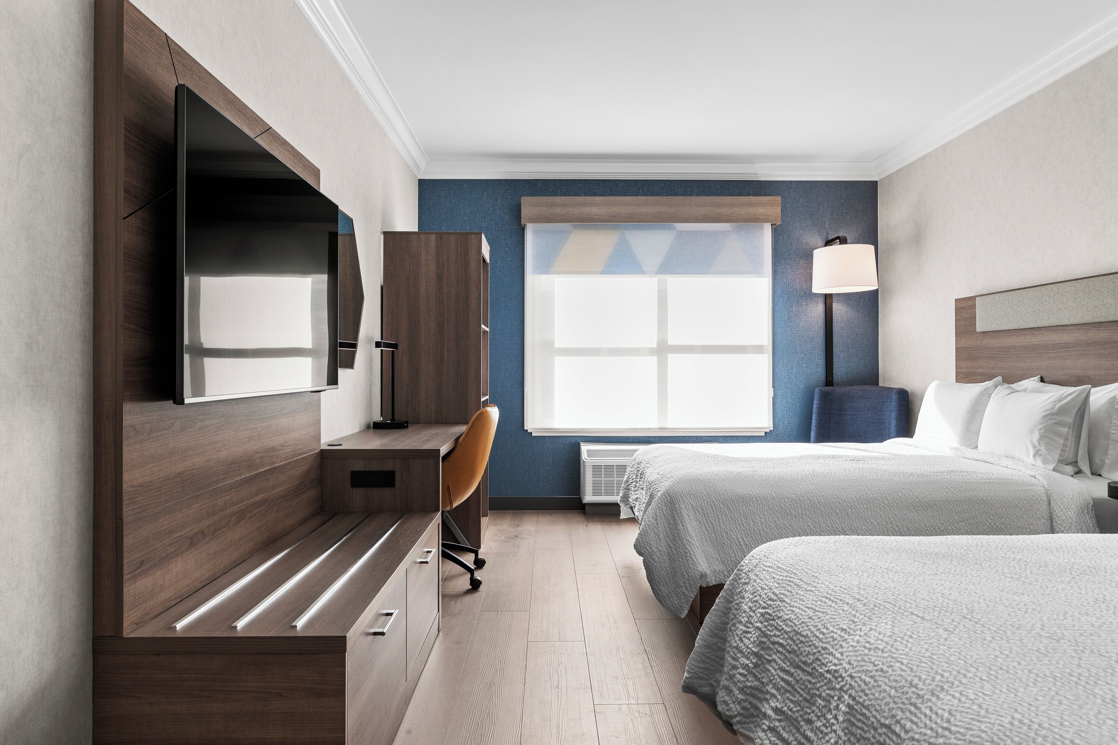 Une chambre d’hôtel avec un bureau en bois, une chaise, un lit recouvert d’une couette blanche, un téléviseur fixé au mur et une fenêtre donnant sur le ciel.