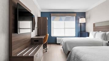 Une chambre d’hôtel avec un bureau en bois, une chaise, un lit recouvert d’une couette blanche, un téléviseur fixé au mur et une fenêtre donnant sur le ciel.